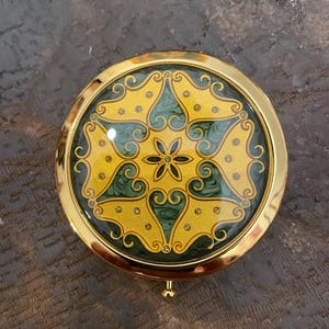 May include: A gold compact mirror with a green and yellow floral design. The mirror has a hinged lid and a small knob on the bottom.