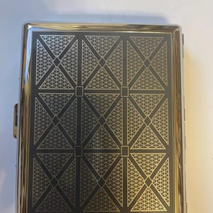 May include: A rectangular, silver-toned cigarette case with a geometric design. The case features a black background with a repeating pattern of gold-colored triangles and squares. The edges of the case are polished and reflective.