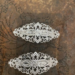 Könnte beinhalten: Zwei silberfarbene Haarspangen mit einem komplizierten, durchbrochenen Filigran-Design. Jede Spange hat eine längliche, ovale Form mit symmetrischen Blumen- und Schnörkelmustern, die ein poliertes Metall-Finish und detaillierte Arbeit zeigen.