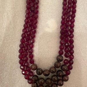 Puede incluir: Un collar de múltiples hilos hecho de cuentas facetadas de color rojo oscuro y cuentas redondas marrones.