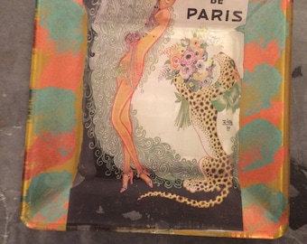 May include: A square glass ashtray with a gold rim and a colorful abstract design. The ashtray features a vintage illustration of a woman in a feather boa and a leopard with a bouquet of flowers. The text "CASINO DE PARIS" is printed on the image.