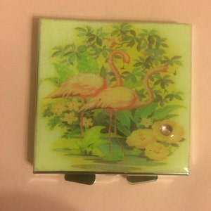 May include: A vintage compact mirror with a hinged lid. The lid features a colourful illustration of two pink flamingos standing in a tropical garden with green leaves and yellow flowers.
