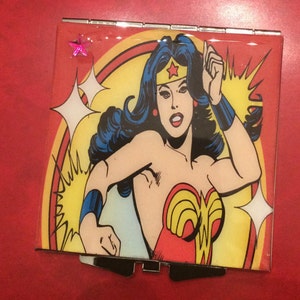 May include: A compact mirror with a colorful image of Wonder Woman in a red, yellow, and blue costume. The background is yellow with a red star on the top left corner.