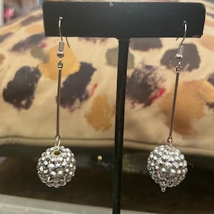 May include: A pair of silver-toned earrings. Each earring features a long, thin, straight bar suspended from a fish hook. At the end of each bar is a round, sparkling, crystal-like ball. The earrings are displayed against a patterned background.