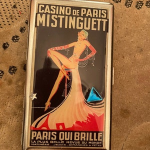 May include: A silver cigarette case with a colorful image of a woman in a red dress and the text "Casino de Paris Mistinguett Paris Qui Brille La Plus Belle Revue Du Monde Matinees Jeudis Dimanches & Fetes".