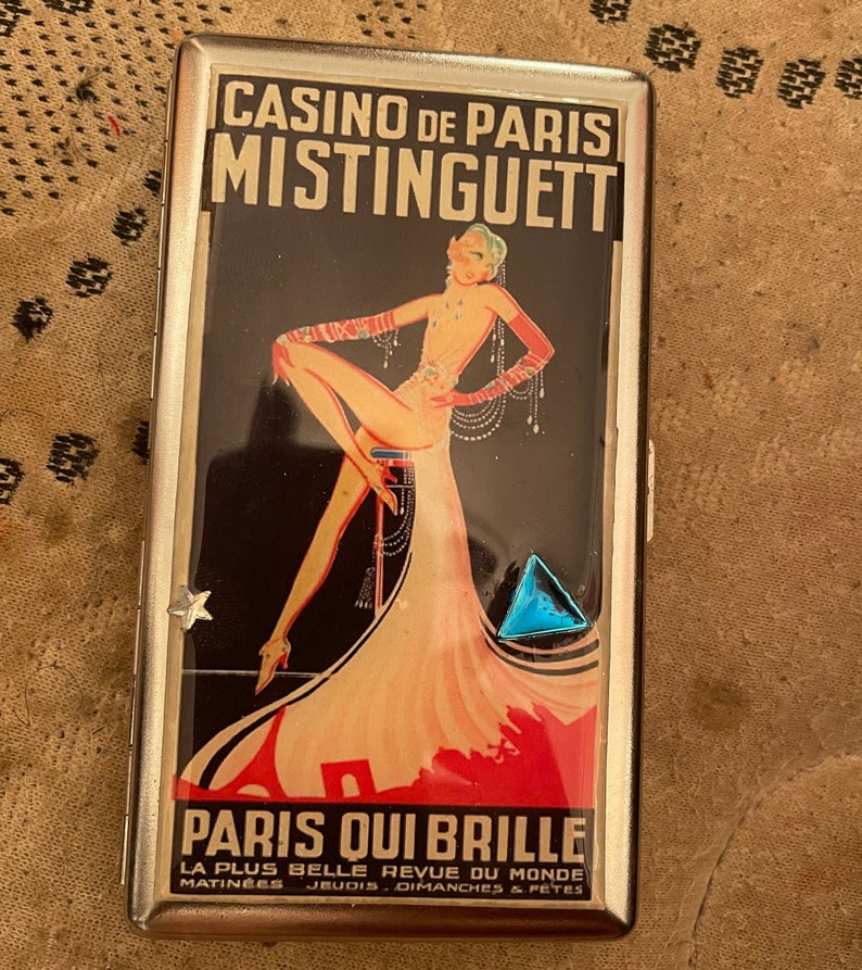 May include: A silver cigarette case with a colorful image of a woman in a red dress and the text "Casino de Paris Mistinguett Paris Qui Brille La Plus Belle Revue Du Monde Matinees Jeudis Dimanches & Fetes".