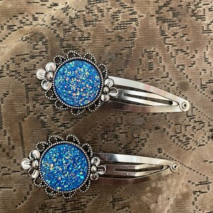 以下が含まれることがあります： 2つのシルバーヘアクリップ。各クリップは、シルバーの花柄のデザインに囲まれた、丸く青いキラキラしたセンターを備えています。クリップは、テクスチャ加工された茶色の背景に配置されています。 各クリップは約6cmです。