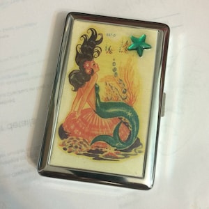 May include: A silver cigarette case with a mermaid illustration. The mermaid has long black hair, a green tail, and is sitting on a seashell. The case has a green star charm attached to it. The text "887-D" is printed on the case.