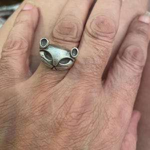 Può includere: Un anello argentato con un design stilizzato a forma di volto di gatto. L'anello presenta una superficie dettagliata e strutturata con evidenti dettagli di occhi e orecchie. L'anello a forma di gatto è indossato su un dito, mostrando il design del gioiello.