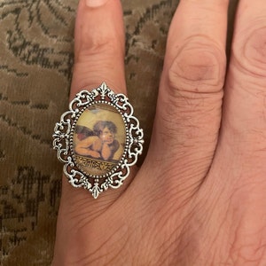 Puede incluir: Un anillo de plata con un camafeo de forma ovalada que representa a un querub&iacute;n con alas. El camafeo est&aacute; engastado en un marco de filigrana con un dise&ntilde;o detallado.