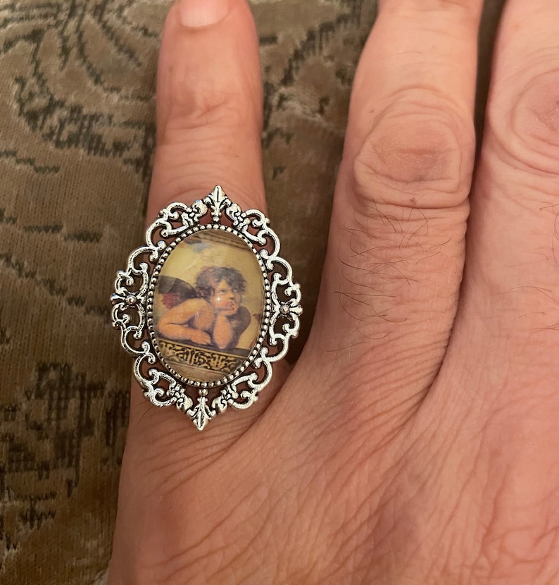 Puede incluir: Un anillo de plata con un camafeo de forma ovalada que representa a un querub&iacute;n con alas. El camafeo est&aacute; engastado en un marco de filigrana con un dise&ntilde;o detallado.