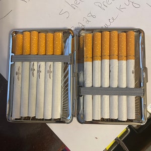 Pu&ograve; includere: Un porta sigarette d'argento con un elastico nero che tiene un pacchetto di sigarette. Il porta sigarette &egrave; aperto e le sigarette sono visibili. Il porta sigarette &egrave; su una superficie bianca.