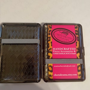 May include: A black metal cigarette case with a cheetah print design. The case is open and shows a pink card with the text "Cheetahrama by Deborah Gregory" and "Handcrafted Fierce Accessories & Cheetahlicious Finds". The card also includes the email address "mamacheetahs@aol.com" and the website "cheetahrama.etsy.com".