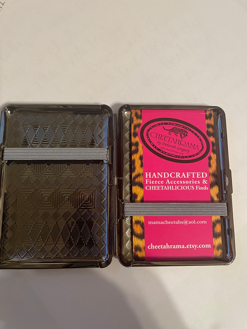 May include: A black metal cigarette case with a cheetah print design. The case is open and shows a pink card with the text "Cheetahrama by Deborah Gregory" and "Handcrafted Fierce Accessories & Cheetahlicious Finds". The card also includes the email address "mamacheetahs@aol.com" and the website "cheetahrama.etsy.com".