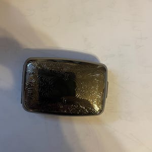 Puede incluir: Un estuche rectangular de metal gris oscuro con un intrincado diseño floral en espiral en la tapa. El estuche tiene una superficie brillante y reflectante y un área oscura y rectangular en el centro.