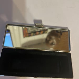 May include: A silver compact mirror with a black velvet interior. The mirror is closed and the lid is slightly ajar.