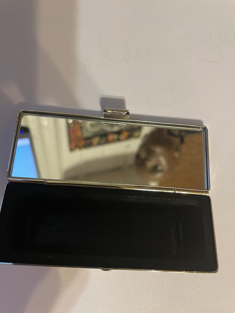 May include: A silver compact mirror with a black velvet interior. The mirror is closed and the lid is slightly ajar.