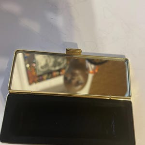 May include: A gold-colored compact mirror with a black velvet interior. The mirror is open and the reflection of a person is visible.
