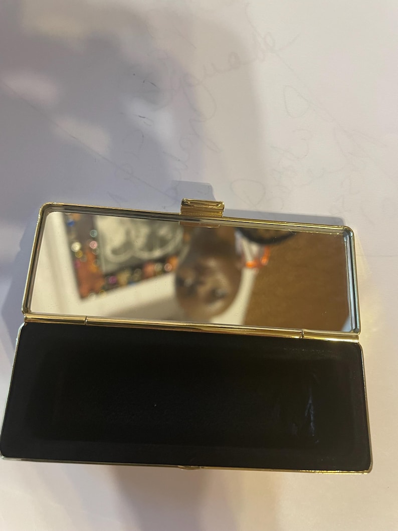 May include: A gold-colored compact mirror with a black velvet interior. The mirror is open and the reflection of a person is visible.