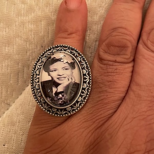 May include: A silver ring with an oval-shaped glass cameo featuring a black and white photo of a woman with short hair. The cameo is surrounded by a decorative silver frame.