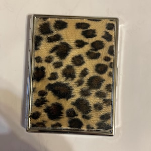 May include: A rectangular cigarette case with a silver metal frame and a leopard print design on the front. The leopard print features brown spots on a tan background. The case is closed and appears to be new.