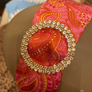 Puede incluir: Una diadema de color rosa y naranja con estampado de paisley y una gran hebilla redonda dorada con pedrería.