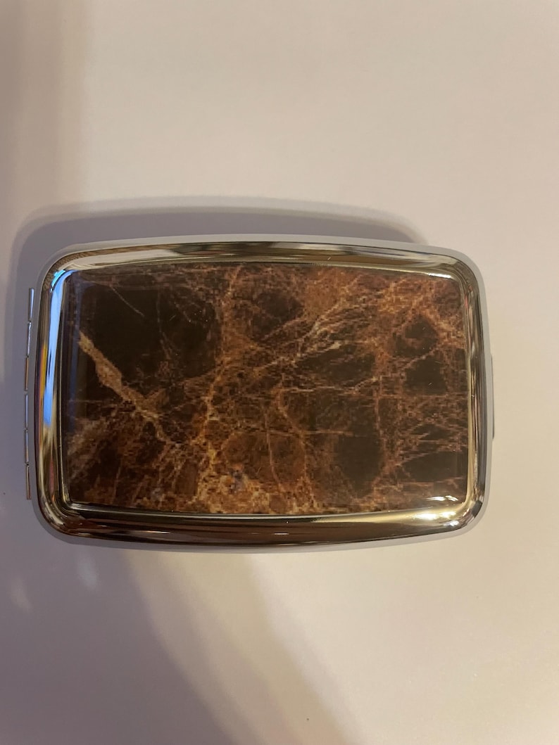 May include: A rectangular metal case featuring a polished silver-tone frame and a dark brown faux marble inlay on the top surface. The case has rounded corners and a smooth, reflective finish, showcasing a detailed veined pattern in shades of brown.