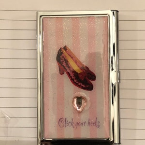 Può includere: Un porta carte argentato con uno sfondo di glitter rosa e bianco. L'immagine presenta un paio di scarpette di rubino rosse con un cuore rosa e il testo "Click your heels".