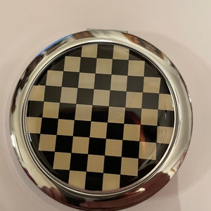 Può includere: Uno specchio compatto rotondo di colore argento con un design a scacchi bianco e nero sul davanti.