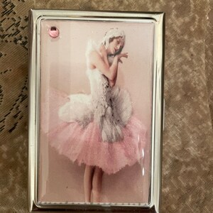 Puede incluir: Una caja de metal plateada con una imagen de bailarina rosa y blanca en la parte delantera. La bailarina lleva un tutú blanco y una corona blanca. La caja tiene un strass rosa en la parte superior.