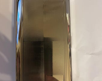Könnte beinhalten: Eine rechteckige, goldfarbene Zigarettenetui mit strukturierter Oberfläche. Das Etui hat ein geometrisches Design mit horizontalen und vertikalen Linien und einen kleinen, rechteckigen Akzent. Das Etui ist geschlossen und hat abgerundete Ecken.