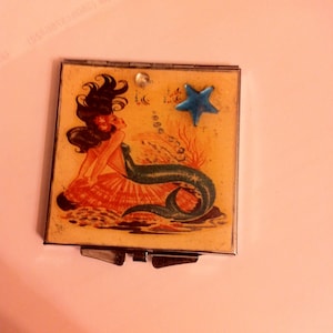 May include: A vintage compact mirror with a mermaid illustration. The mermaid is depicted with long dark hair, a blue tail, and a starfish on the top of the mirror.