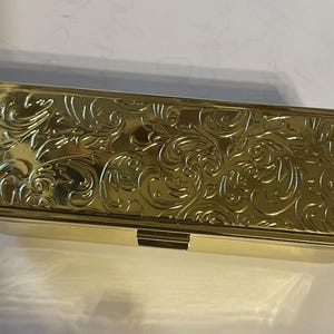 May include: A gold-colored rectangular box with an ornate, embossed floral design. The box has a hinged lid and a decorative clasp.