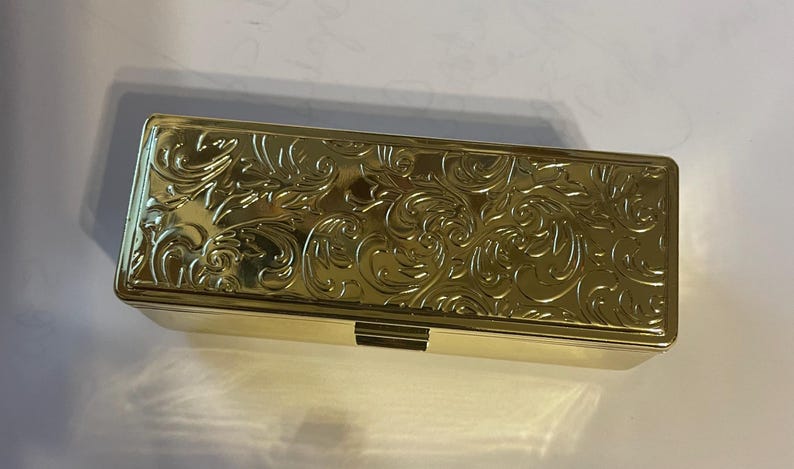 May include: A gold-colored rectangular box with an ornate, embossed floral design. The box has a hinged lid and a decorative clasp.