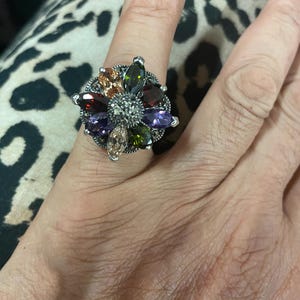 Può includere: Un anello d'argento con un design a forma di fiore con una varietà di pietre preziose colorate, tra cui verde, viola e marrone.