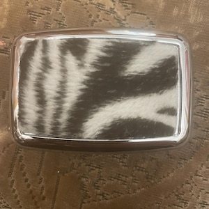 Puede incluir: Una hebilla de cinturón rectangular con un marco de metal plateado pulido que rodea una incrustación central de piel con estampado animal en blanco y negro. El diseño tiene esquinas redondeadas y un borde metálico liso y reflectante.
