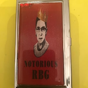 May include: A silver metal card holder with a red background featuring a portrait of a woman wearing a crown and a black and white patterned collar. The text "NOTORIOUS RBG" is printed in black on the red background.