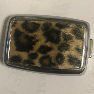 May include: A rectangular belt buckle featuring a polished silver-tone metal frame surrounding a central inlay of faux fur with a brown and black leopard print pattern. The buckle has a smooth, reflective border and a functional metal attachment loop.