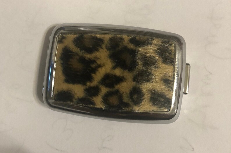 May include: A rectangular belt buckle featuring a polished silver-tone metal frame surrounding a central inlay of faux fur with a brown and black leopard print pattern. The buckle has a smooth, reflective border and a functional metal attachment loop.