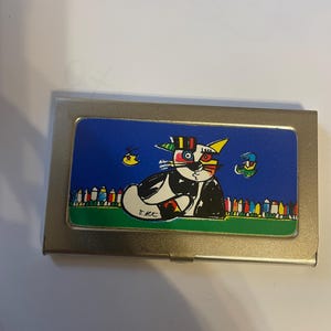 Könnte beinhalten: Ein silberner Visitenkartenhalter mit einem farbenfrohen Cartoon-Katzen-Design. Die Katze ist schwarz und weiß mit einem gelben Auge und einem bunten Hut. Der Hintergrund ist blau mit zwei kleinen Vögeln und einer grünen Wiese.