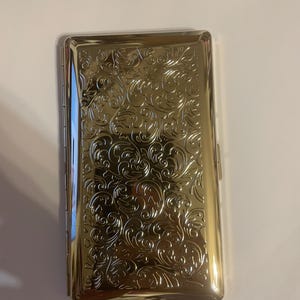 May include: A gold-colored cigarette case with an ornate, embossed floral design. The rectangular case has rounded edges and a hinged closure. The surface reflects light, highlighting the intricate patterns of the embossed design.