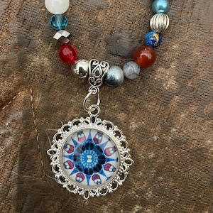 Peut inclure: Un collier de perles avec un pendentif en argent. Le pendentif a un cadre rond et orné et un motif floral bleu et rouge. Le collier comporte une variété de perles colorées, dont blanches, bleues, rouges et argentées.