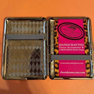 May include: A silver metal cigarette case with a cheetah print design. The case is open and the inside is lined with a silver metal pattern. The case has a black elastic band on the outside and a pink label with the text "CHEETAHRAMA by Deborah Gregory" and "HANDCRAFTED Fierce Accessories & CHEETAHLICIOUS Finds" and the website address "mamacheetahs@aol.com" and "cheetahrama.etsy.com".