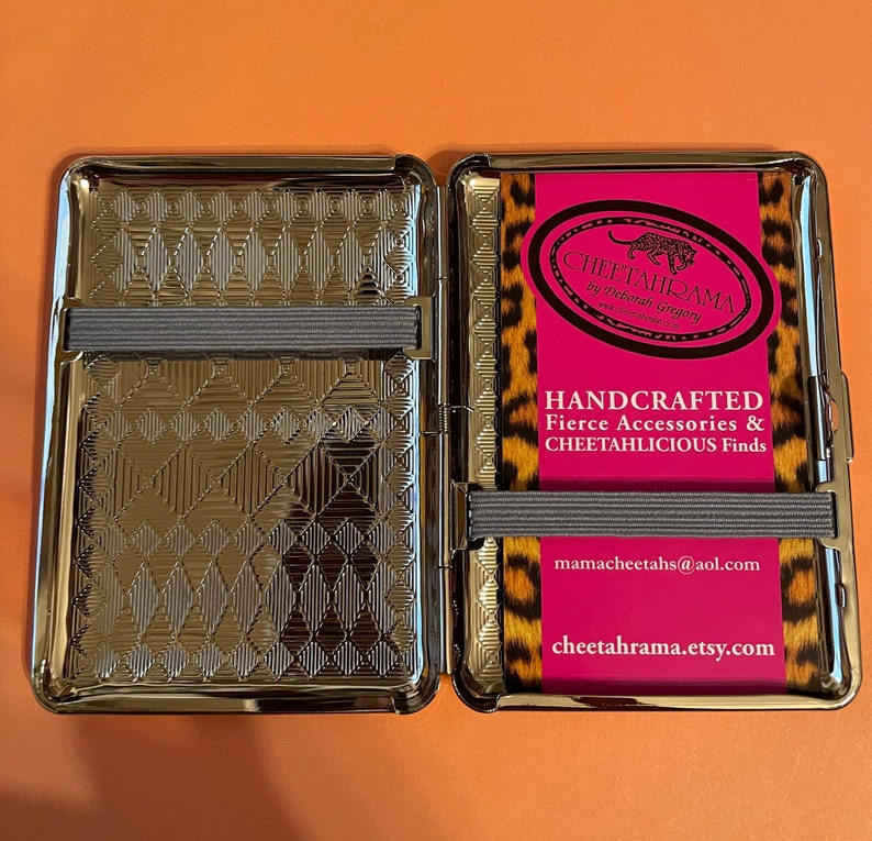 May include: A silver metal cigarette case with a cheetah print design. The case is open and the inside is lined with a silver metal pattern. The case has a black elastic band on the outside and a pink label with the text "CHEETAHRAMA by Deborah Gregory" and "HANDCRAFTED Fierce Accessories & CHEETAHLICIOUS Finds" and the website address "mamacheetahs@aol.com" and "cheetahrama.etsy.com".