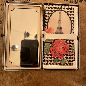 May include: A silver cigarette case with a mirror on one side and a black and white houndstooth pattern with a red rose and a picture of the Eiffel Tower on the other side.