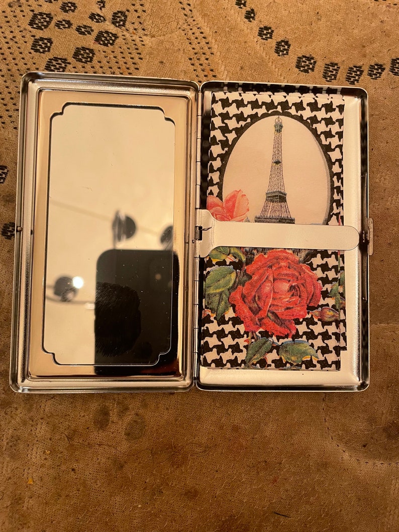May include: A silver cigarette case with a mirror on one side and a black and white houndstooth pattern with a red rose and a picture of the Eiffel Tower on the other side.