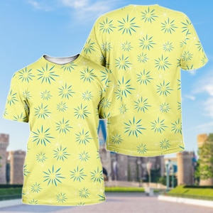 May include: A light yellow t-shirt featuring a repeating pattern of blue starbursts and white geometric lines. The shirt has a crew neck and short sleeves. The background shows a blurred image of a castle and blue sky.