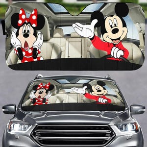 May include: Car sunshade featuring Mickey Mouse and Minnie Mouse. Mickey is in a red shirt, waving from the driver's seat. Minnie is in a red dress with white polka dots. The sunshade is black with a car interior background.