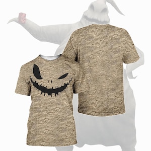 May include: A tan t-shirt featuring a black, grinning face design. The shirt has a textured pattern and a crew neck. A second shirt is visible, with a white silhouette of a character in the background. The design is reminiscent of a Halloween theme.