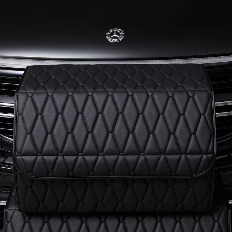 K&ouml;nnte beinhalten: Schwarzes, gestepptes Leder-Autozubeh&ouml;r mit Rautenmuster. Das Zubeh&ouml;r befindet sich an der Vorderseite eines schwarzen Autos, im Hintergrund ist das Mercedes-Benz-Logo zu sehen.